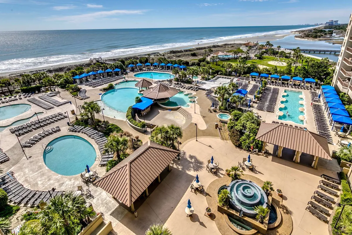 A wide exterior shot showcases the resort’s sprawling waterpark oasis, complete with sparkling swimming pools surrounded by palm trees, lounge chairs, and cabana structures. The setting is lively yet relaxing, inviting guests to enjoy sunbathing, swimming, and poolside socializing with views of the beachfront in the distance.