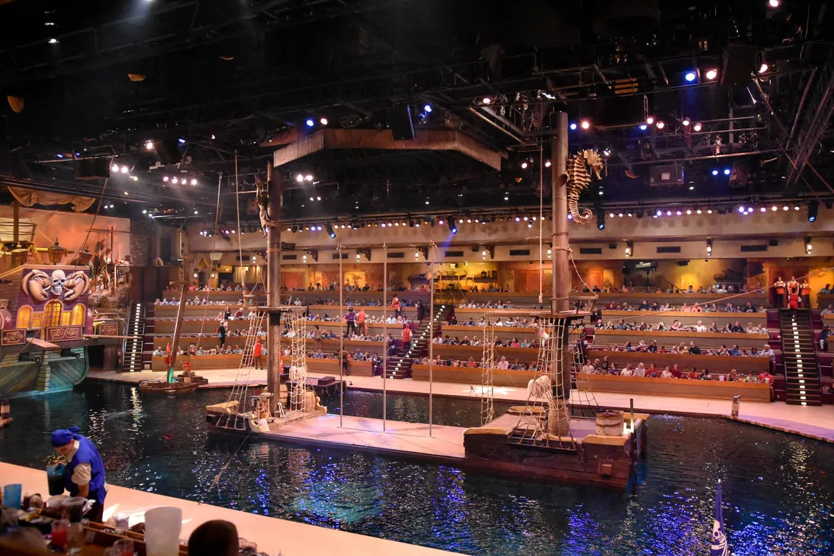 An interior shot of a performance venue set with a water stage, themed decor, audience seating, and performers in action. The atmosphere is energetic and immersive, hinting at live shows and family entertainment.
