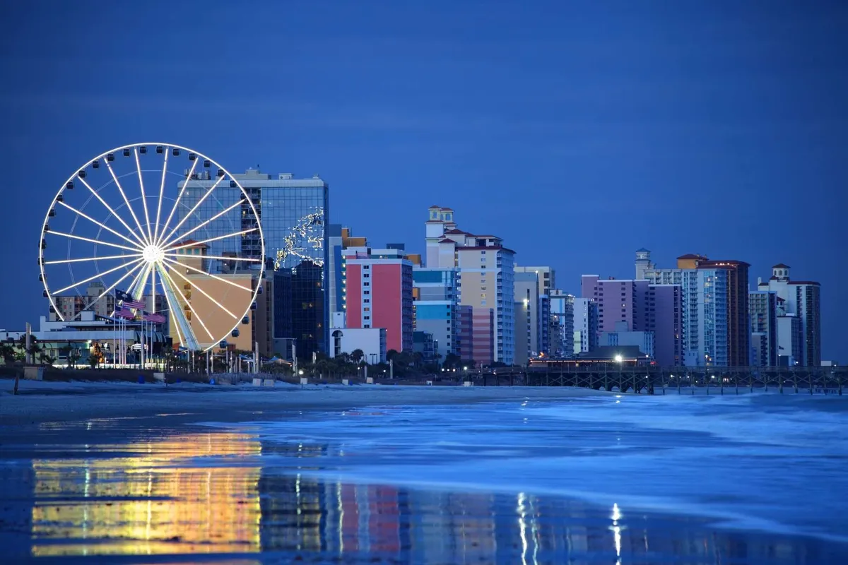 A picturesque nighttime view featuring a brightly lit Ferris wheel, colorful waterfront buildings, and their reflections sparkling on the calm water. The scene evokes the excitement of amusement park rides and seaside entertainment, perfect for a family night out.