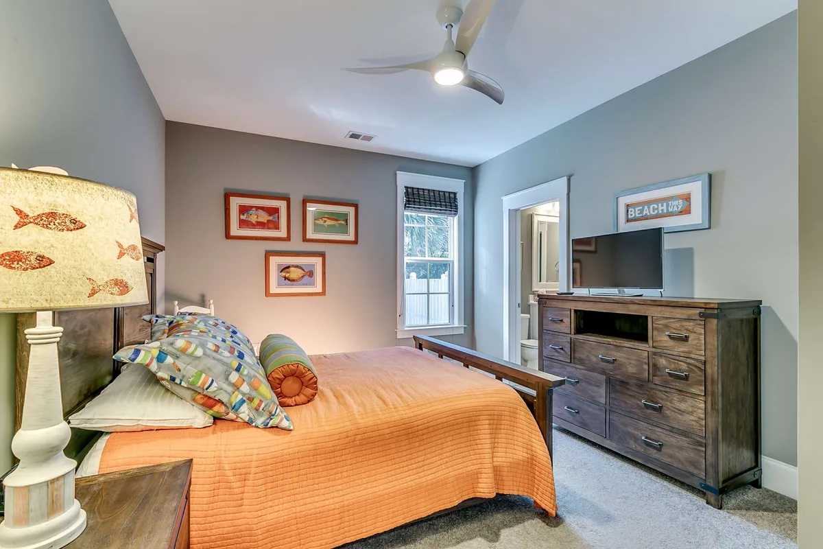 This bedroom features a bed with a vibrant orange quilt and colorful pillows, a fish-themed lamp, wall artwork, and a television unit. The space feels welcoming and relaxed, with plenty of natural light and thoughtful decorative touches that foster a restful environment.