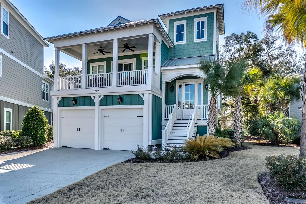 The image showcases the inviting exterior of a single-family house in North Myrtle Beach, with a charming front porch, lush landscaped yard, and stately palm trees. The home's green facade is accented by double garage doors, and the porch glows in the warm light of sunset, offering a peaceful, tropical retreat.