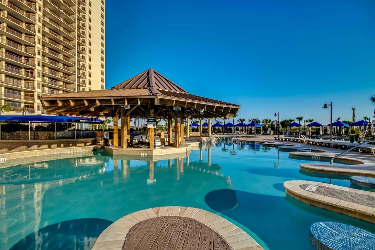 A lively resort pool scene featuring a large swimming pool surrounded by palm trees, a thatched gazebo, plenty of lounge chairs, and a clear blue sky. The setting is inviting for both families and groups, suggesting a variety of activities from swimming and sunbathing to poolside dining and games.
