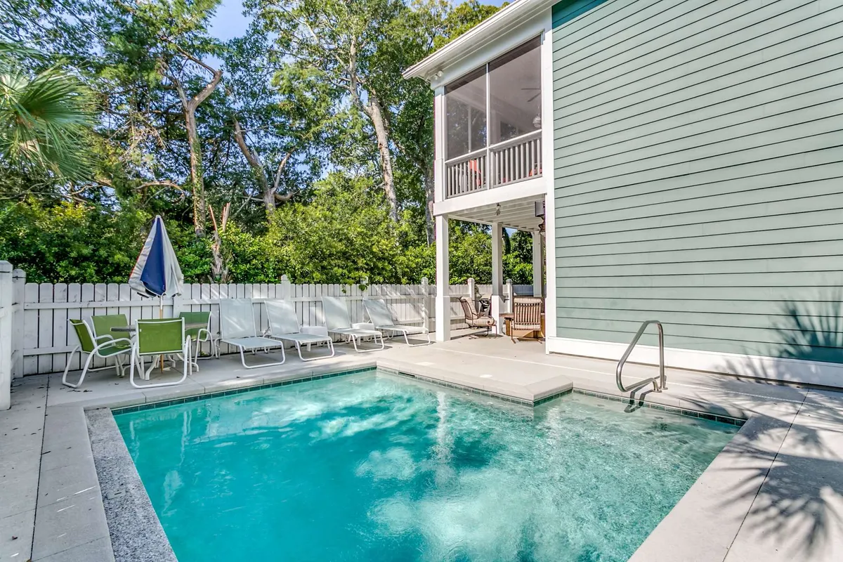 A serene exterior view of a private swimming pool surrounded by lounge chairs and lush greenery. To the right, a screened porch overlooks the pool deck, offering a shaded and breezy retreat. The scene captures a sense of privacy and relaxation, perfect for both daytime swims and sunset lounging.