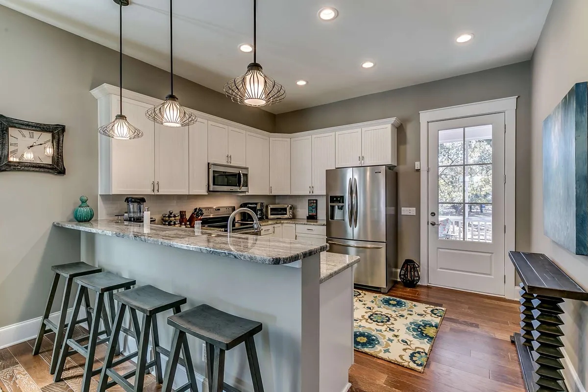 A bright, cheerful kitchen with a large island, hanging light fixtures, stainless steel appliances, and a decorative rug. Natural light pours through the window, emphasizing a space ideal for cooking and socializing.