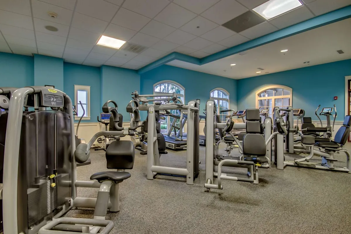 A modern, light-filled fitness center features rows of state-of-the-art exercise machines against bright blue walls and large windows. The scene is energetic, with individuals engaged in workouts, personal training, and group fitness activities, emphasizing a motivating environment.